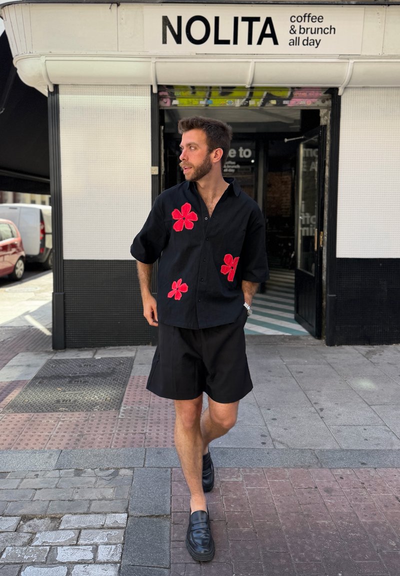Homme portant une chemise noire à fleurs rouges et un short noir marchant devant un café nommé Nolita proposant café et brunch toute la journée.