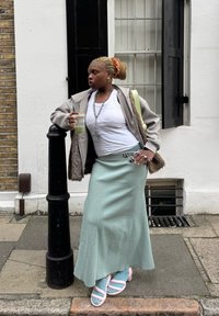 Világosszürke, túlméretezett kabát szőrmés részletekkel, fehér póló, menta zöld szoknya és rózsaszín pántos szandálok. Zöld italt tart a kezében. Városi környezet.