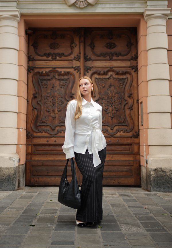 Mujer con blusa blanca y falda negra a rayas, que lleva un bolso negro, está de pie en una calle adoquinada frente a unas grandes puertas dobles ornamentadas de madera.