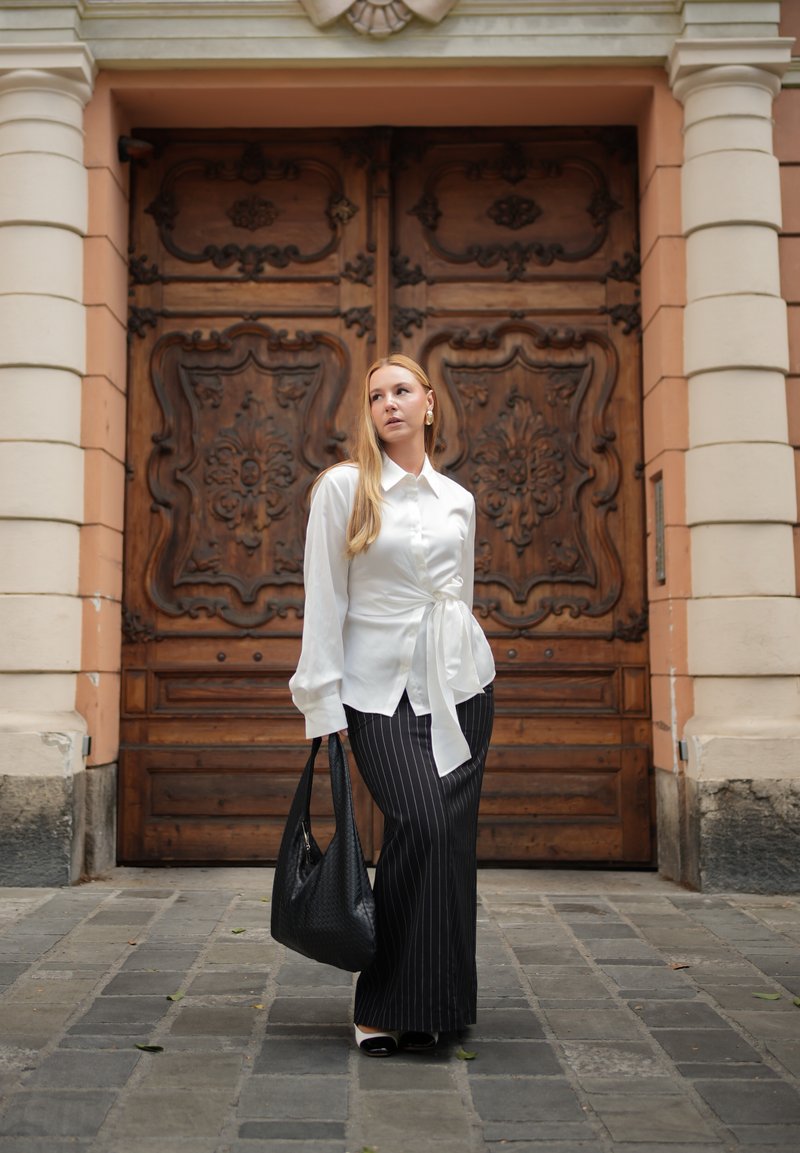 Mujer con blusa blanca y falda negra a rayas, que lleva un bolso negro, está de pie en una calle adoquinada frente a unas grandes puertas dobles ornamentadas de madera.