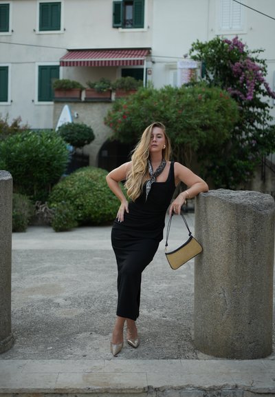 Vestido negro ajustado con escote redondeado, combinado con tacones metálicos de punta y un pequeño bolso beige. Los accesorios incluyen una bufanda de estampado.
