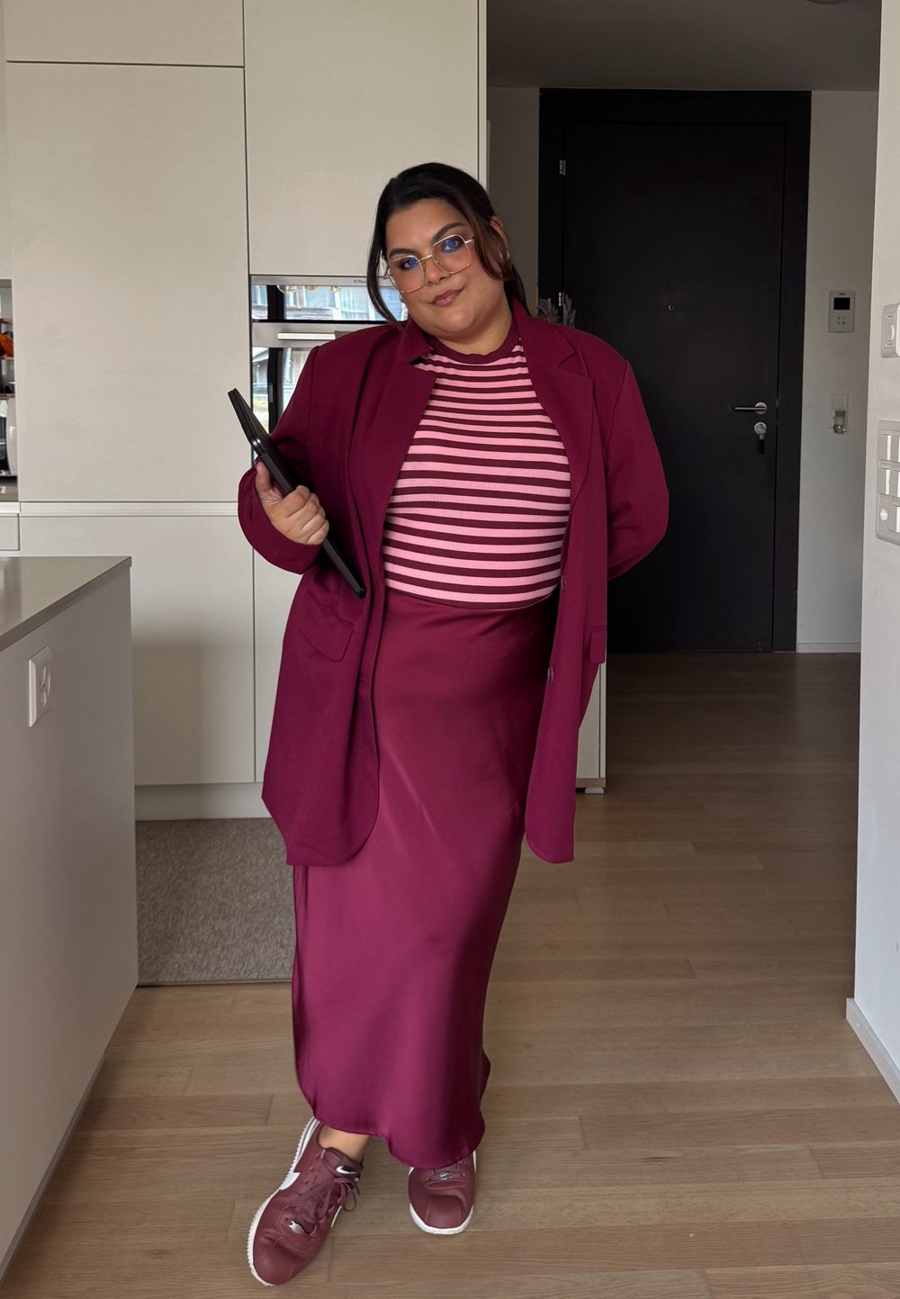 Burgundin bleiseri vaaleanpunaisella ja valkoisella raidallisella paidalla, yhdistettynä saman sävyiseen pitkään hameeseen ja burgundin värisiin tennareihin, pitäen mustaa tablettia.