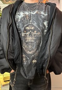 Svart puffjacka med dragkedjan öppen, vilket visar en mörk t-shirt med dödskallemotiv, lager av kedjor och smala svarta jeans med kedjedetaljer.