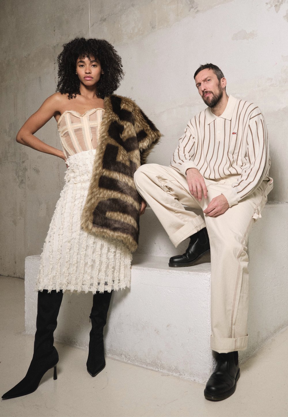 Top de corpete bege, saia branca com franjas texturizadas, botas de salto alto pretas. Homem com pullover riscado, calças bege, botas de tornozelo pretas. Acessório de braço em pelo.