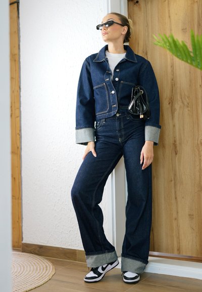 Chaqueta de mezclilla y jeans de talle alto en lavado oscuro con puños claros, combinados con gafas de sol negras y un pequeño bolso negro. Zapatillas negras y blancas.