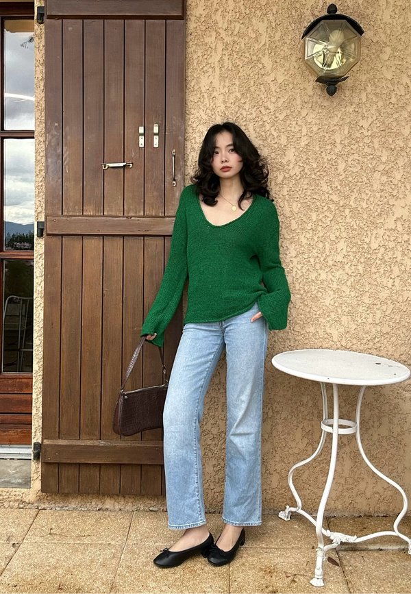Une femme en pull vert, en jean clair et en chaussures plates noires pose près d’une porte en bois. Elle tient un sac à main brun et se trouve à proximité d’une table de terrasse blanche.
