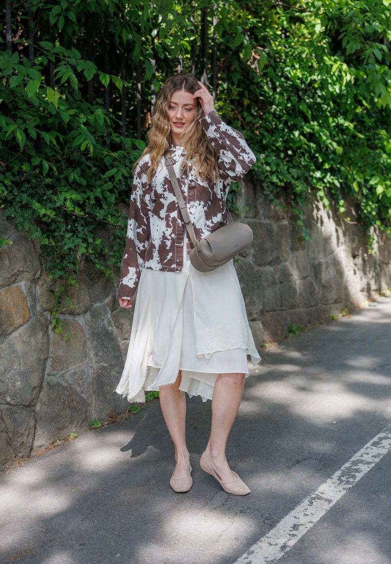 Femme aux longs cheveux ondulés porte une veste marron et blanche à motifs, une jupe blanche fluide, des ballerines beiges et un sac à bandoulière gris, debout près d'un mur en pierre.