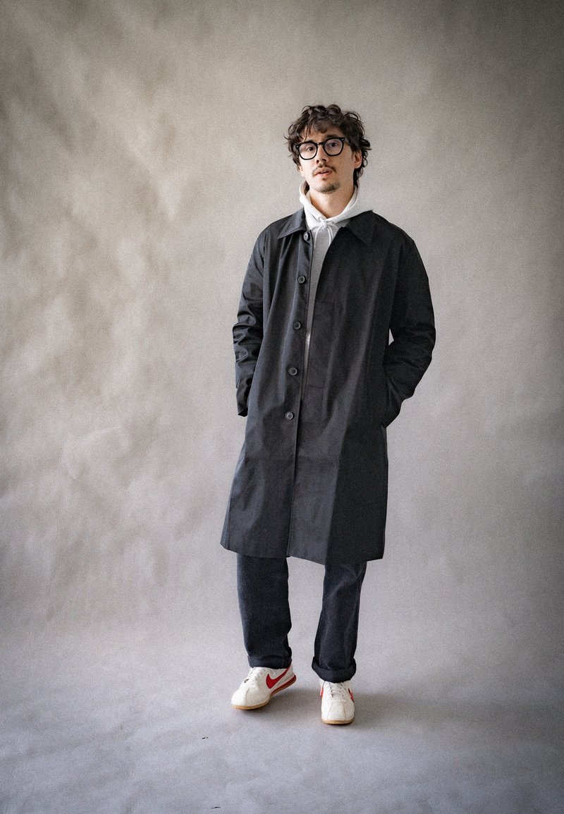 Cappotto lungo nero con chiusura a bottoni, colletto a punta, maniche lunghe, indossato sopra un hoodie bianco e jeans scuri. Sneakers bianche con dettagli rossi.
