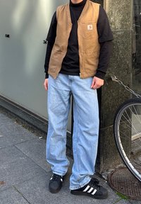 Gilet marrone sopra una maglietta nera a maniche lunghe, jeans azzurri chiari e scarpe nere con strisce bianche. In piedi accanto a una bicicletta.