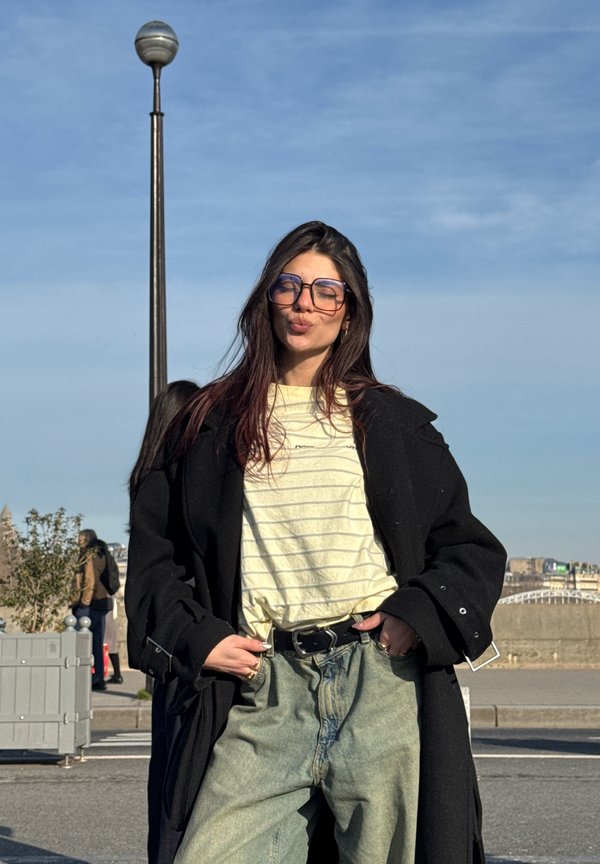 Junge Frau mit Brille, die einen schwarzen Mantel, ein gestreiftes Hemd und Jeans trägt, posiert im Freien unter klarem blauem Himmel in der Nähe einer Straßenlaterne.