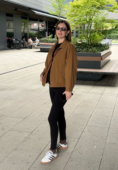 Chaqueta marrón con cintura elástica, top negro, leggins negros y zapatillas blancas con rayas negras; de pie al aire libre en un entorno de plaza.