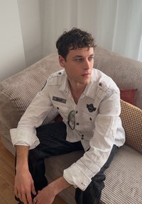 Jeune homme aux cheveux bouclés et foncés, portant une chemise blanche à boutons, assis pensivement sur un canapé beige avec des coussins à motifs.