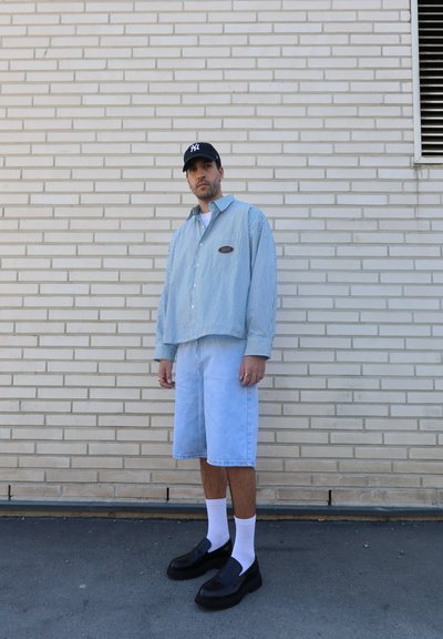 Camisa corta de rayas azul y blanca, pantalones cortos de mezclilla claros, zapatos negros tipo slip-on y calcetines blancos, frente a un fondo de pared de ladrillo claro.