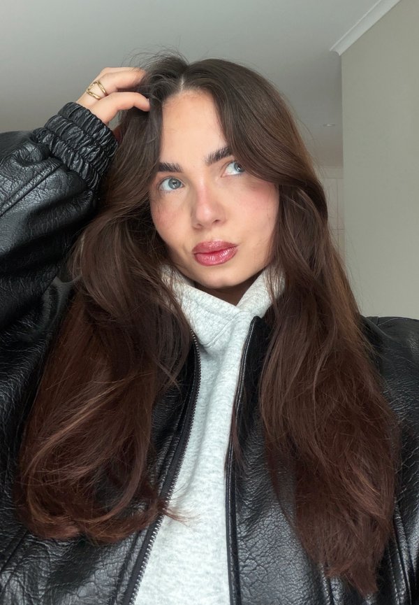 Mujer con cabello largo y castaño, con chaqueta de cuero negra y sudadera gris, tocándose el cabello con una mano, mirando ligeramente hacia arriba y hacia un lado.