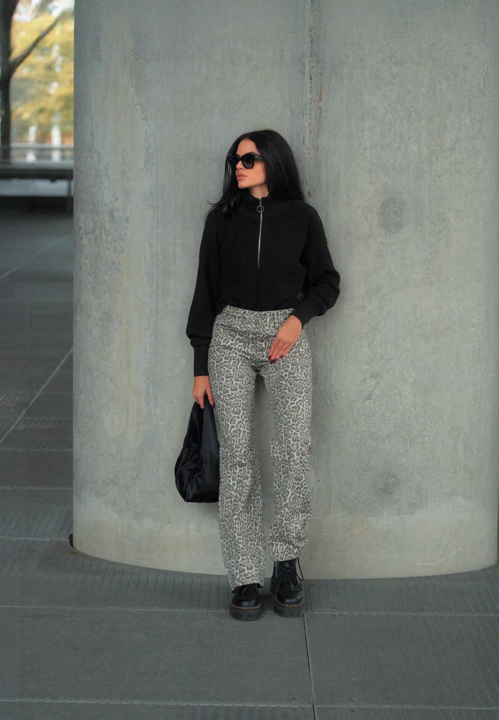 Μαύρο ζακέτα με φερμουάρ, παντελόνι λεοπάρ, μαύρες πλατφόρμες και μία μαύρη τσάντα. Στηρίζεται σε έναν τοίχο από σκυρόδεμα.