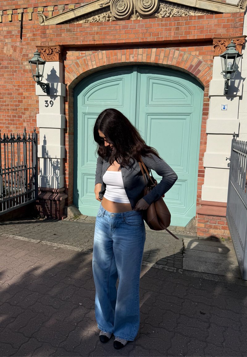 Femme portant un haut blanc, un cardigan gris, un jean à jambes larges et des lunettes de soleil, debout devant un mur en briques avec une grande porte vert menthe.