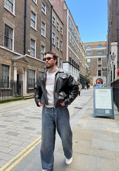 Hombre con gafas de sol, chaqueta de cuero negra y vaqueros caminando por una calle estrecha de la ciudad bordeada de edificios de ladrillo y un cartel de restaurante.