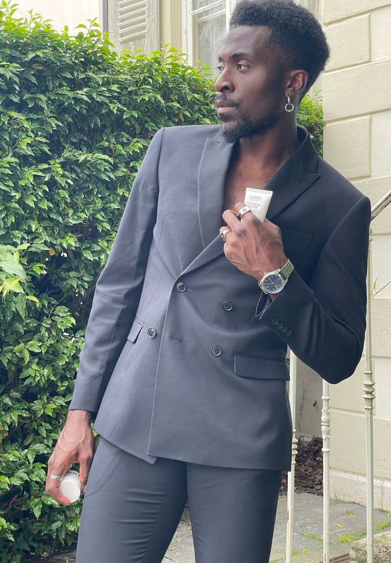 Blazer noir à double boutonnage en tissu lisse, associé à un pantalon ajusté assorti. Tient un produit en tube blanc, se tenant à côté de verdure.