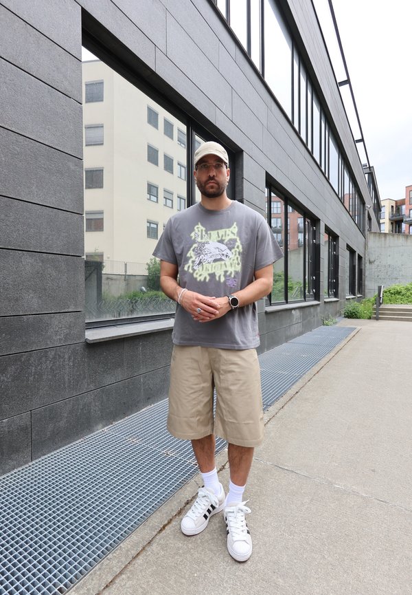 Homme portant un short beige, un t-shirt gris avec un motif graphique, des baskets blanches et une casquette, debout sur le trottoir près d’un bâtiment moderne avec de grandes fenêtres.