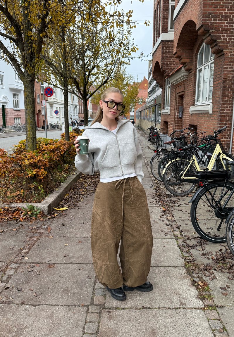 Felpa con zip grigio chiaro, pantaloni marroni a gamba larga e scarpe nere. Tiene in mano un bicchiere verde da bevanda. Contesto urbano con biciclette e alberi.