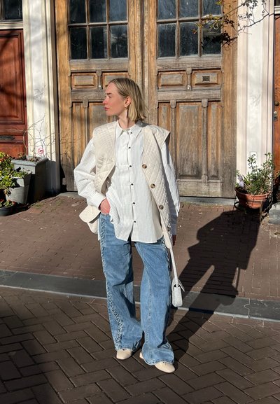 Chaleco acolchado blanco sobre una camisa de rayas de botones y vaqueros de pierna ancha en azul claro con detalles laterales. Zapatos planos beige y bolso pequeño.