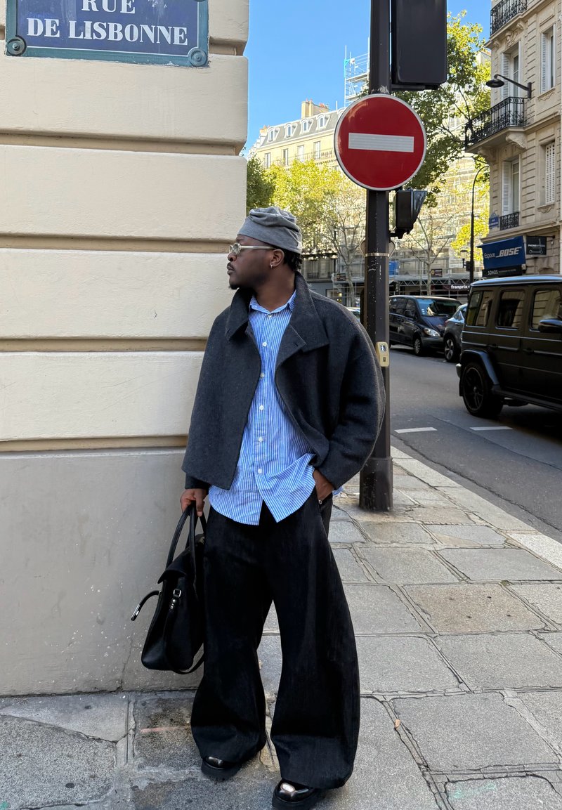 Veste noire surdimensionnée portée sur une chemise rayée bleu et blanc, associée à un pantalon large noir et un bonnet gris ; tenant un sac noir.
