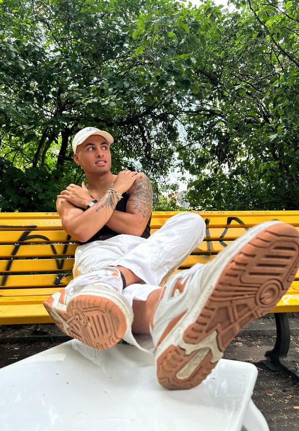 Zapatillas deportivas blancas con acentos naranjas, usadas con pantalones blancos; persona sentada en un banco amarillo bajo un follaje verde.