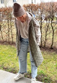 Μαλακό, ελαιόχρωμο γιλέκο πάνω από ένα ανοιχτό γκρί πουλόβερ, συνδυασμένο με μπλε τζιν και άσπρα αθλητικά παπούτσια. Το φόντο περιλαμβάνει καφετιά θάμνους και πράσινο γρασίδι.