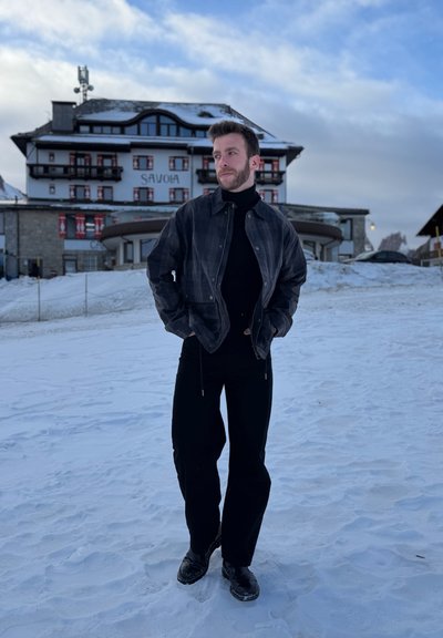 Hombre con chaqueta y pantalones oscuros de pie sobre la nieve frente a un gran edificio alpino con la inscripción "SAVOIA" bajo un cielo nublado.