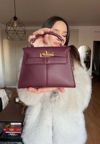 Femme en manteau de fourrure blanche tenant un petit sac à main en cuir bordeaux avec un fermoir doré dans un salon moderne.