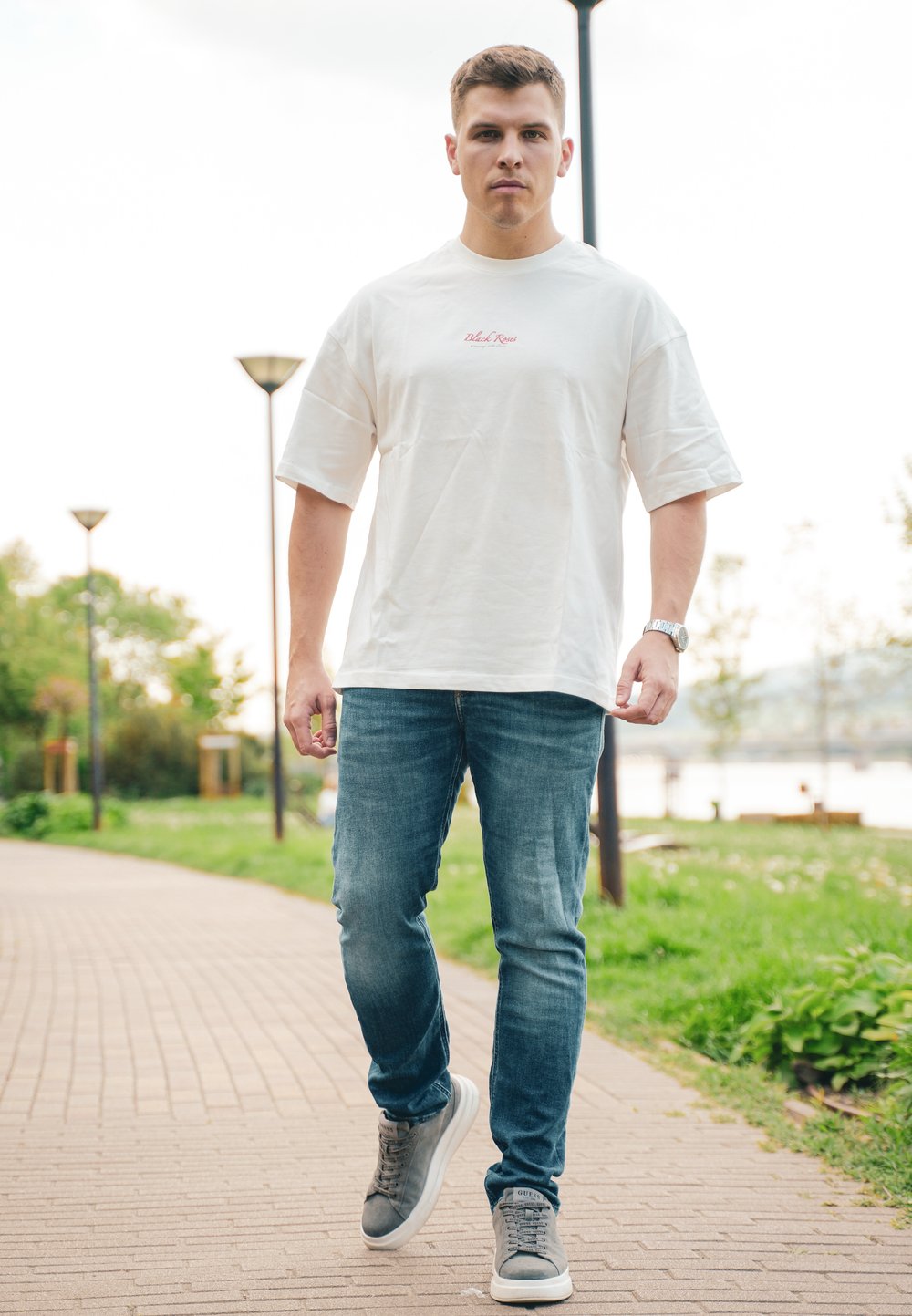 Vit t-shirt med kort ärm och röd text, blå jeans och grå sneakers. Scenen visar en gångväg omgiven av grönska och lampstolpar.