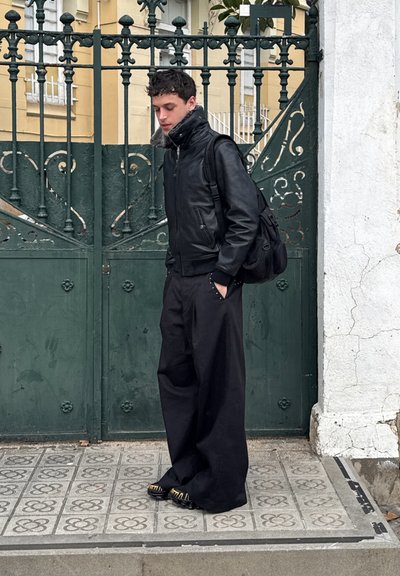 Joven con chaqueta de cuero negra, pantalones anchos negros y mochila, de pie sobre una acera con patrón frente a una puerta verde de metal ornamentada.