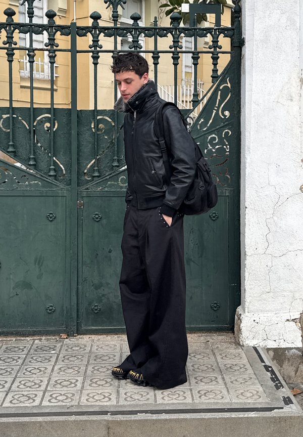 Joven con chaqueta de cuero negra, pantalones anchos negros y mochila, de pie sobre una acera con patrón frente a una puerta verde de metal ornamentada.