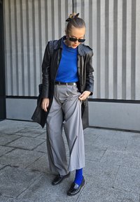 Μαύρο δερμάτινο παλτό, μπλε πουλόβερ, γκρι παντελόνι με φαρδιά εφαρμογή, αντιθετικά μπλε καλτσάκια, μαύρες μοκέτες, υφασμάτινο φόντο με κατακόρυφες ρίγες.