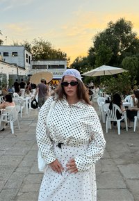 Fehér felső fekete pöttyökkel, bő szabású, gombos, hozzáillő fehér szoknyával párosítva. Kiegészítők között fekete napszemüveg és fényes fejkendő található.