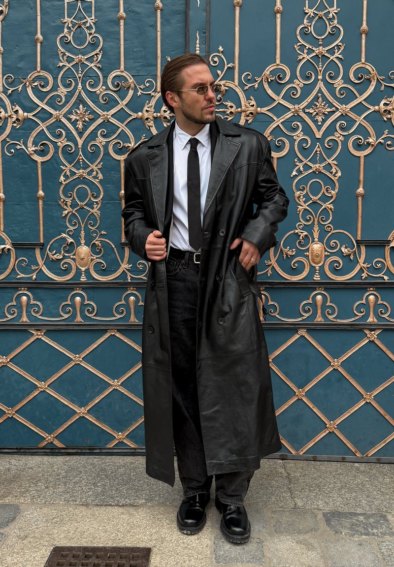 Homme portant un long manteau en cuir noir, une chemise blanche, une cravate noire, des lunettes de soleil et des bottes, se tenant devant une porte en fer forgé bleue et dorée ornée.