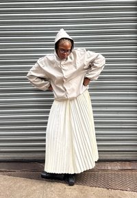 Beige gewatteerde jas met een gladde textuur, gedragen over een witte geplooide rok. De outfit wordt gecombineerd met zwarte schoenen op een gestructureerd oppervlak.