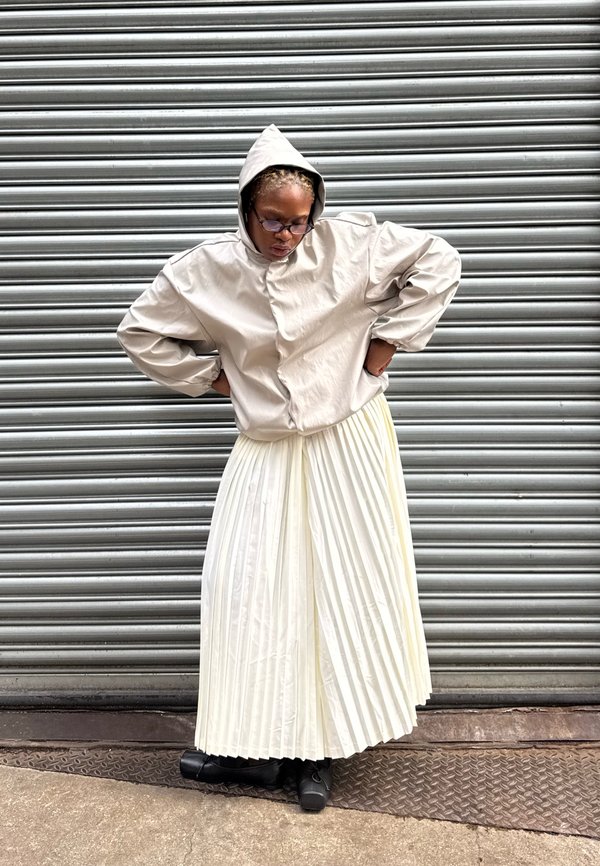 Veste à capuche beige à texture lisse, portée sur une jupe plissée blanche. La tenue est assortie à des chaussures noires sur une surface texturée.