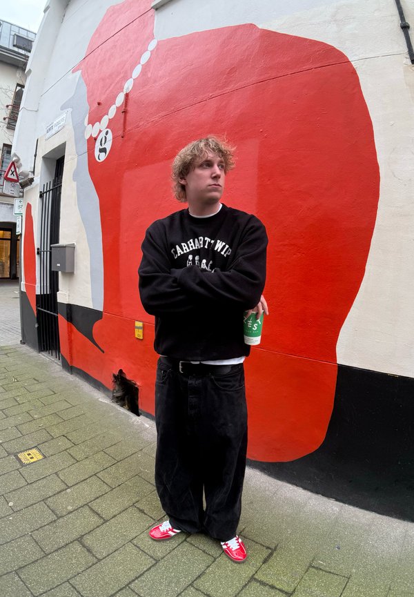 Svart överdimensionerad sweatshirt med vit text, lösa svarta jeans, röda sneakers, håller en grön mugg, framför en stor röd mural på en vägg.