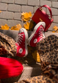 Adidași înalți din pânză roșie, cu talpă de cauciuc albă și sfoară. Pantaloni bej cu imprimeu leopard, pulover tricotat roșu și o geantă roșie în apropiere.