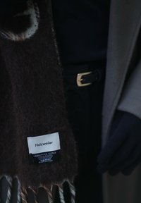 Cachecol de mistura de lã de cordeiro e alpaca castanho escuro com uma textura suave, apresentando um rótulo e bordas frisadas. Usado com uma luva preta.