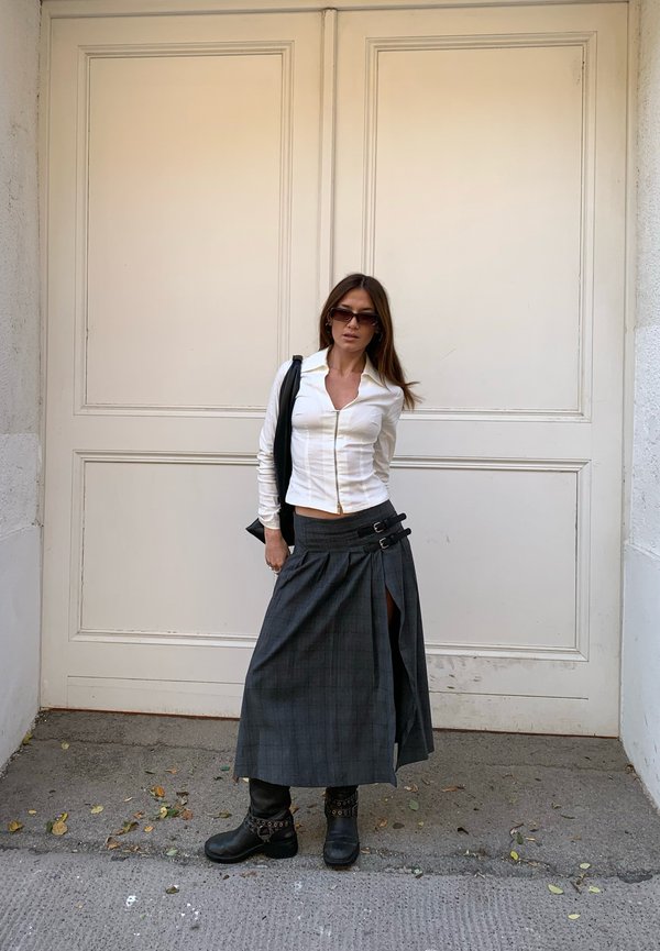 Blusa blanca ajustada con cremallera, falda corta de cuadros gris hasta la rodilla con aberturas laterales y botas negras hasta el tobillo con herrajes decorativos. Bolso negro.