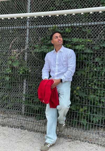 Camisa de lino blanca de botones, jeans de color azul claro, zapatillas marrones con rayas blancas, sosteniendo un suéter rojo, de pie cerca de una cerca de cadena.