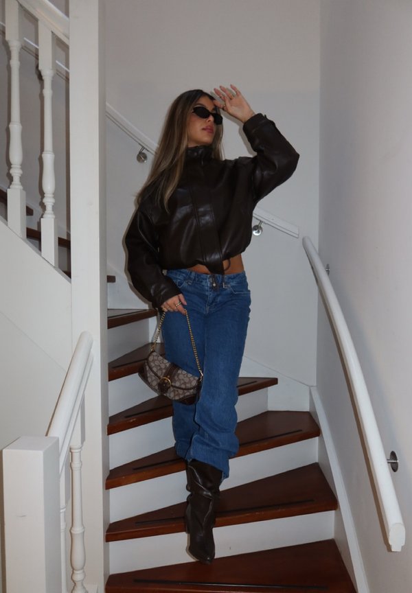 Femme portant une veste en cuir noire, des lunettes de soleil, un jean bleu et des bottes, descendant des escaliers en bois en tenant un petit sac à chaîne.