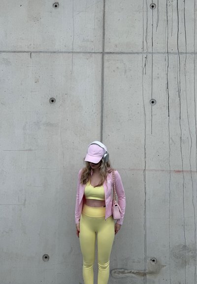 Mujer con ropa deportiva amarilla, chaqueta rosa, gorra y auriculares, de pie contra una pared de concreto, con la cabeza baja y las manos a los lados.