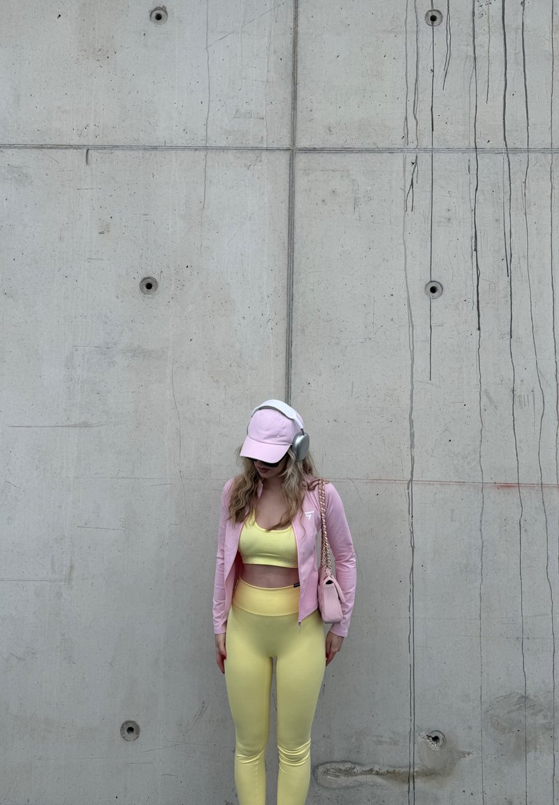 Femme vêtue d'une tenue de sport jaune et d'une veste rose, portant une casquette et un casque, debout contre un mur en béton, la tête baissée et les mains le long du corps.