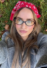 Sivi vuneni kaput s teksturiranim završetkom, duga svijetlosmeđa kosa, okrugle naočale i crvena bandana s bijelim uzorcima na pozadini trave.