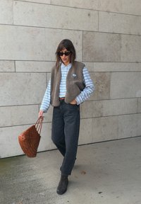 Brūns vilnas bezmuguras veste virs gaiši zila svītrainā garajose krekla, tumši džinsi un brūni potīšu zābaki. Tur rokām austu brūnu somu.