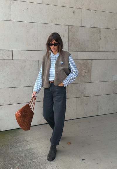 Chaleco de forro marrón sobre una camisa de rayas azul claro de manga larga, jeans oscuros y botas marrones de tobillo. Sostiene una bolsa tote de mimbre marrón.
