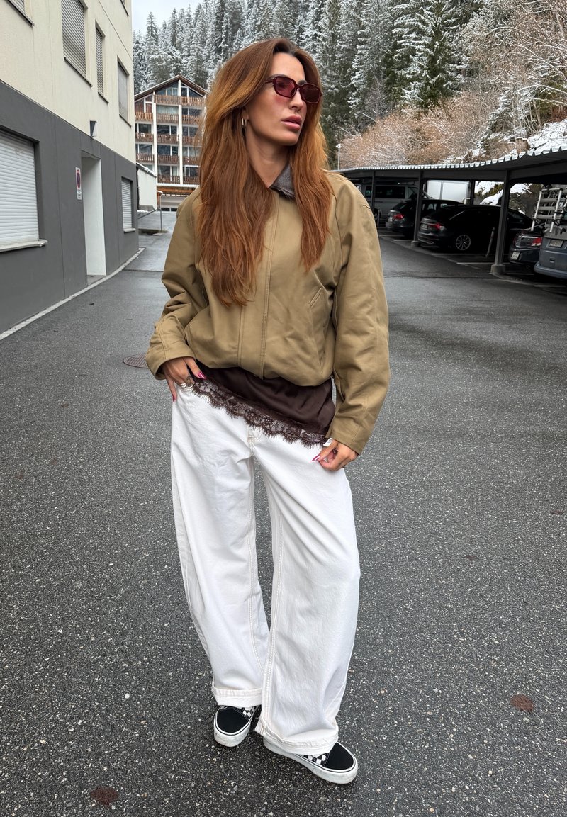 Kvinde med langt rødligt hår iført en beige jakke, solbriller, brun skjorte med blondekant, brede hvide bukser og sorte sneakers på en snedækket parkeringsplads på bjerget.